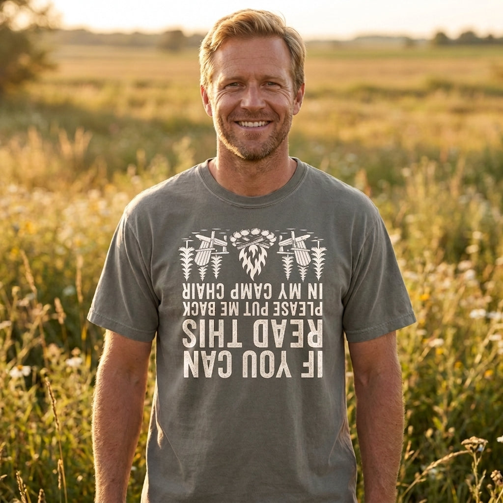 Smiling man in a dark gray If You Can Read This camp chair t-shirt with white text and bee illustrations, standing in a sunlit grassy field.