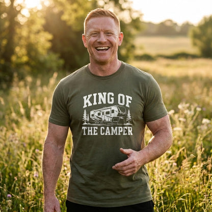 A smiling man standing outdoors in a grassy field at sunset, wearing an olive green graphic T-shirt with the text 'KING OF THE CAMPER' and an RV illustration.