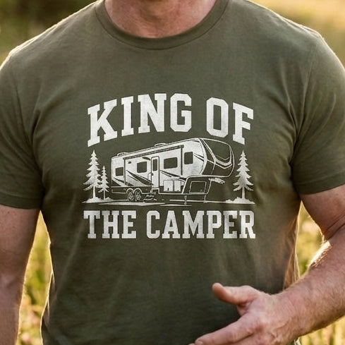 A smiling man standing outdoors in a grassy field at sunset, wearing an olive green graphic T-shirt with the text 'KING OF THE CAMPER' and an RV illustration.