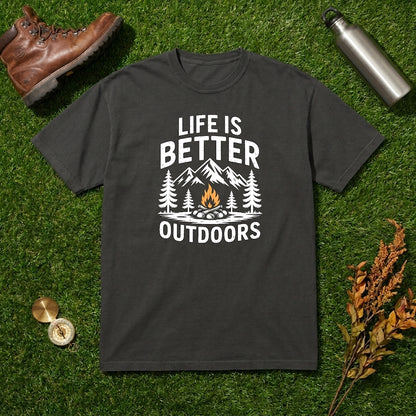 A dark charcoal gray t-shirt with a white graphic reading 'LIFE IS BETTER OUTDOORS' and showing mountains, trees, and a campfire. The shirt is laid flat on green grass with outdoor props including a brown leather boot, a silver water bottle, and dried flowers.