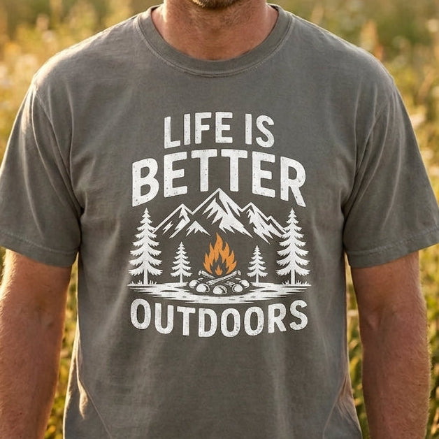 A smiling man standing in a sunlit field wearing a gray graphic T-shirt with the phrase 'LIFE IS BETTER OUTDOORS' and a mountain, trees, and campfire illustration.