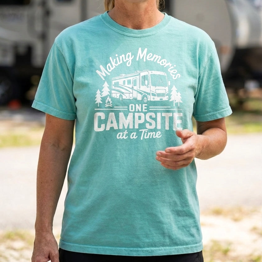 A woman wearing a teal graphic t-shirt with a white print that reads 'Making Memories One Campsite at a Time' with an illustration of a camper van and trees, standing outdoors in front of a blurred RV and trees.