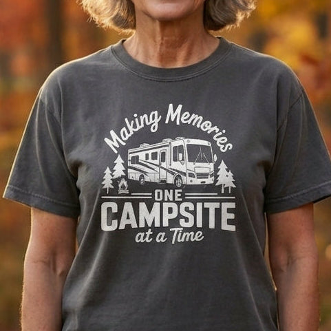A smiling older woman stands outdoors in a forest during autumn, wearing a dark gray graphic T-shirt that reads 'Making Memories at the Campsite' with a small RV illustration.