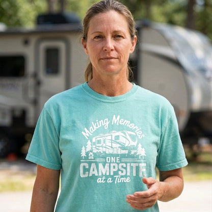 A woman wearing a mint green graphic t-shirt that reads 'Making Memories One Campsitra at a Time' with a white camper graphic, standing outdoors in front of a blurred campsite trailer.