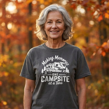 A smiling woman with gray hair stands outdoors in a forest with autumn foliage, wearing a dark gray graphic T-shirt that reads 'Making Memories One Campsite at a time' with a camper illustration.