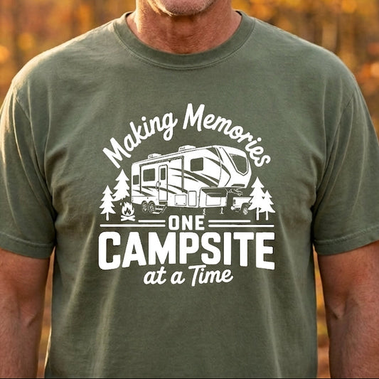 A smiling man wearing an olive green graphic t-shirt with a white camping RV design and the text 'Making Memories ONE CAMPSITE at a Time', set against a warm, blurred autumn forest background.