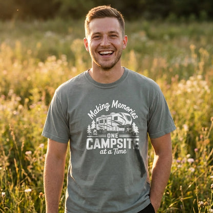 A smiling man standing outdoors in a grassy field at sunset, wearing a gray graphic T-shirt with the text 'Making Memories One Campsite at a Time' and a camper illustration.