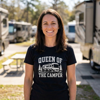 A smiling woman stands outdoors at a campground wearing a black short-sleeve T-shirt with a white graphic that reads 'QUEEN OF THE CAMPER' and shows an RV between trees.