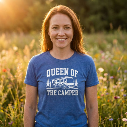 A smiling woman with red hair stands outdoors in a sunlit field wearing a heather blue graphic T-shirt that reads 'QUEEN OF THE CAMPER' with a white RV and trees print.