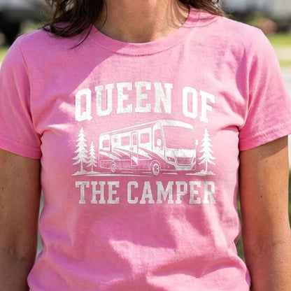 A smiling woman wearing a pink graphic t-shirt that reads 'QUEEN OF THE CAMPER' with a white camper illustration, standing outdoors in front of blurred RVs at a campsite.