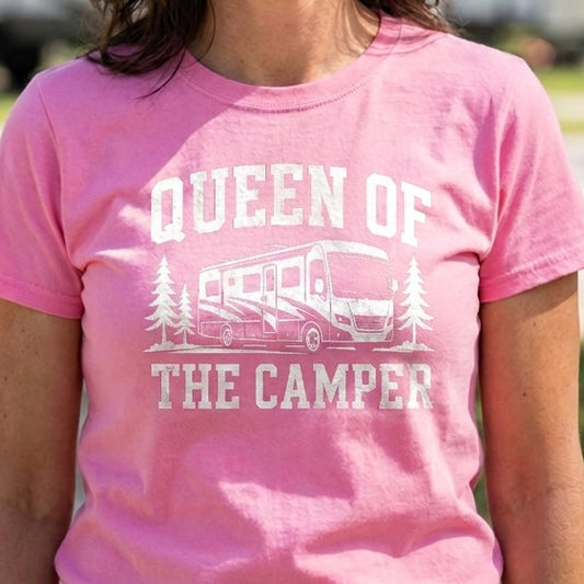 A smiling woman wearing a pink graphic t-shirt that reads 'QUEEN OF THE CAMPER' with a white camper illustration, standing outdoors in front of blurred RVs at a campsite.