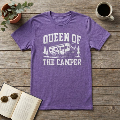 A heather purple short-sleeve t-shirt with a white graphic reading 'QUEEN OF THE CAMPER' and an RV illustration, laid flat on a rustic wood surface with a plant, coffee mug, sunglasses, and open book as props.