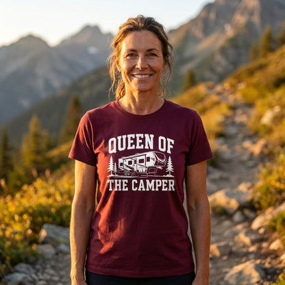 A woman standing outdoors in a mountain landscape wearing a maroon short-sleeve graphic T-shirt with white text reading 'QUEEN OF THE CAMPER' and a small camper illustration.