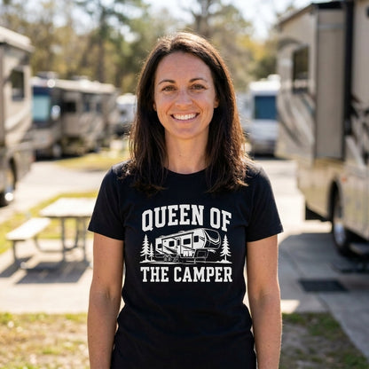 A smiling woman stands outdoors at a campground wearing a black graphic T-shirt that reads 'QUEEN OF THE CAMPER' with a camper illustration. RVs and picnic tables are blurred in the background.