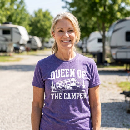 A smiling blonde woman wearing a purple graphic t-shirt that reads 'QUEEN OF THE CAMPER' with an RV and trees, standing outdoors at a campground with RVs in the background.