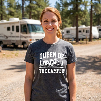 A smiling woman stands outdoors in a campsite setting wearing a dark heather gray graphic t-shirt with the white text 'QUEEN OF THE CAMPER' and a camper illustration. RVs and pine trees are visible in the background.