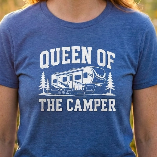 A smiling woman with red hair stands outdoors in a sunlit field wearing a heather blue T-shirt with a white graphic that reads “QUEEN OF THE CAMPER” above and below an RV illustration.