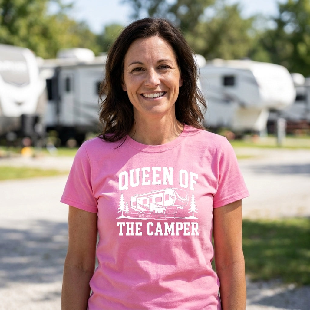 A smiling woman with dark hair stands outdoors in an RV park wearing a pink T-shirt with a white graphic that reads “QUEEN OF THE CAMPER” above and below an RV illustration.
