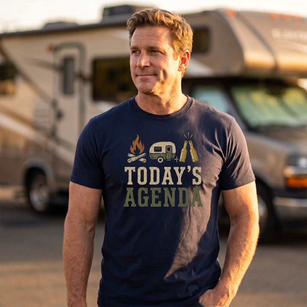 A man wearing a navy blue graphic t-shirt with a camping-themed print that reads 'TODAY'S AGENDA' stands in front of a blurred recreational vehicle.