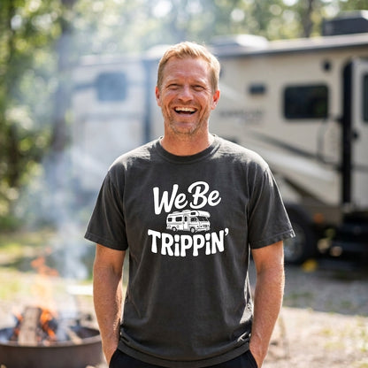 A smiling man wearing a dark charcoal gray t-shirt with a white graphic that reads 'We Be TRIPPIN'' and an RV illustration, standing outdoors in front of a blurred campsite background with an RV and a fire pit.