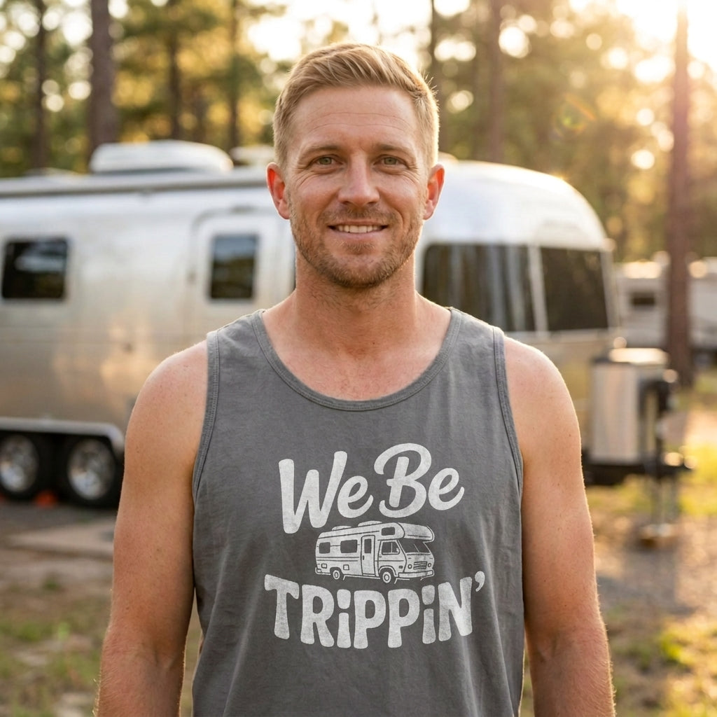 A smiling man wearing a gray tank top with a white graphic that reads 'We Be Trippin'' stands outdoors in front of a silver camper trailer, with trees and warm sunlight in the background.