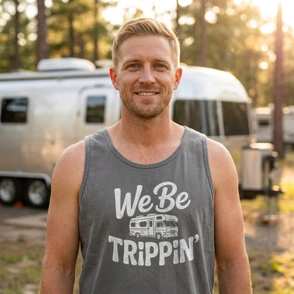 A smiling man wearing a gray tank top with a white graphic that reads 'We Be Trippin'' stands outdoors in front of a silver camper trailer, with trees and warm sunlight in the background.