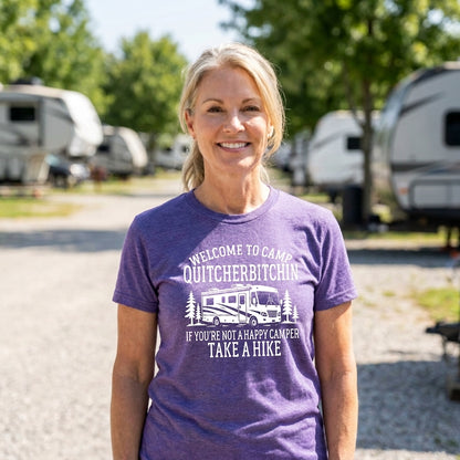 Smiling blonde woman in a purple Welcome to Camp Quitcherbitchin t-shirt with a white RV design, standing at a campground.