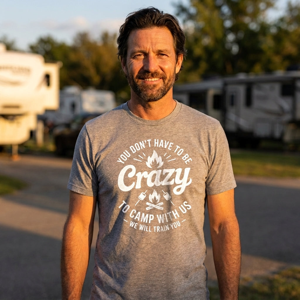 A man wearing a heather gray graphic t-shirt with a white printed design that reads 'WELCOME TO CAMP QUITCHERBITCHIN IF YOU'RE NOT A HAPPY CAMPER TAKE A HIKE'. He is standing outdoors at a campsite with RVs in the background during golden hour.