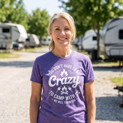 A smiling blonde woman stands outdoors at a campground wearing a heather purple graphic t-shirt that reads 'YOU DON'T HAVE TO BE Crazy TO CAMP WITH US - WE WILL TRAIN YOU'.