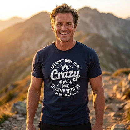 A smiling man standing outdoors in front of mountains at sunset, wearing a navy blue graphic T-shirt with white text that reads, 'YOU DON'T HAVE TO BE Crazy TO CAMP WITH US WE WILL TRAIN YOU.'.