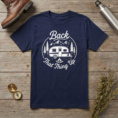 A navy blue short-sleeve t-shirt with a white graphic print reading 'Back That Thing Up' around a camping trailer illustration, laid flat on a rustic wooden surface with a boot, metal water bottle, compass, and dried flowers as props.