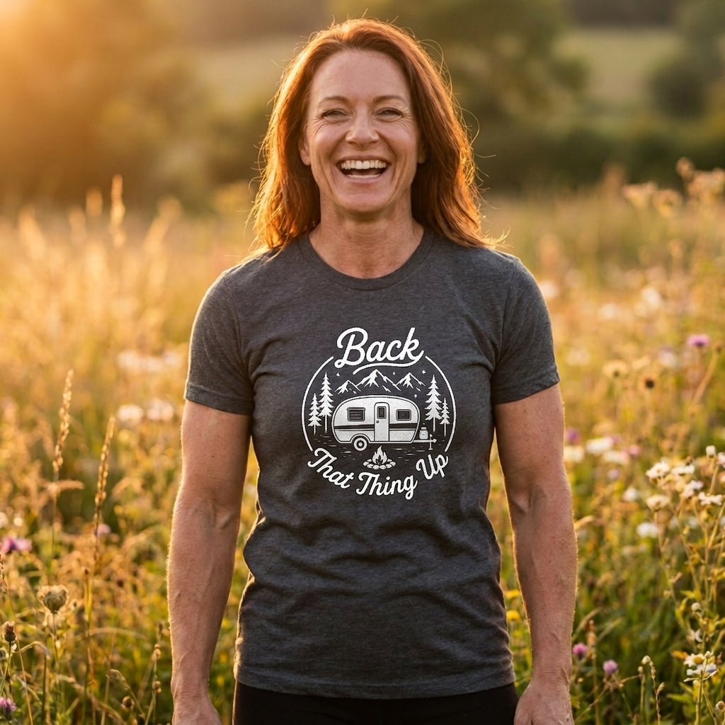 A smiling woman standing outdoors in a sunlit field of wildflowers, wearing a dark heather gray graphic T-shirt with a white camping trailer design and the text 'Back That Thing Up.'.