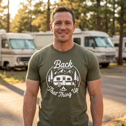 A smiling man wearing an olive green t-shirt with a white graphic that reads 'Back That Thing Up' with a camper van and mountains, standing outdoors in front of campers and trees.