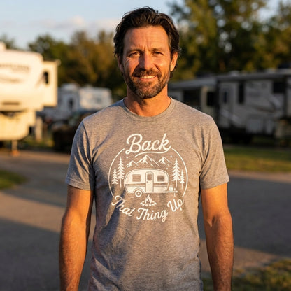 A man wearing a heather gray t-shirt with a white graphic that reads 'Back That Thing Up' with a camping trailer and trees, standing outdoors in front of blurred RVs at sunset.