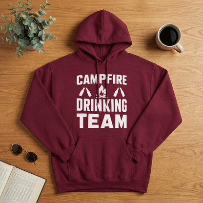 Flat lay of a maroon Campfire Drinking Team hoodie on a wood table with a coffee mug, sunglasses, a plant and an open book