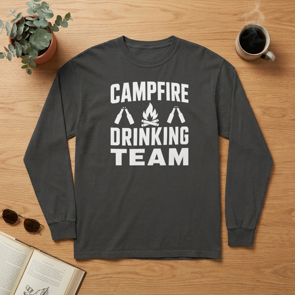 Flat lay of a charcoal gray Campfire Drinking Team long sleeve shirt on a wood table with a coffee mug, sunglasses, a plant and an open book