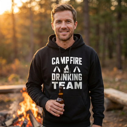 Smiling man in a black Campfire Drinking Team hoodie holding a beer bottle in front of a campfire