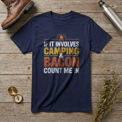 A navy blue graphic t-shirt laid flat on a rustic wooden surface, surrounded by outdoor-themed props including a brown hiking boot, a metal water bottle, a small compass, and dried herbs. The t-shirt features a graphic print that reads 'IF IT INVOLVES CAMPING & BACON COUNT ME IN' with a small campfire icon above.