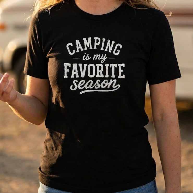 A woman wearing a black graphic t-shirt that reads 'CAMPING is my FAVORITE season' in white lettering, standing outdoors in front of a blurred recreational vehicle.