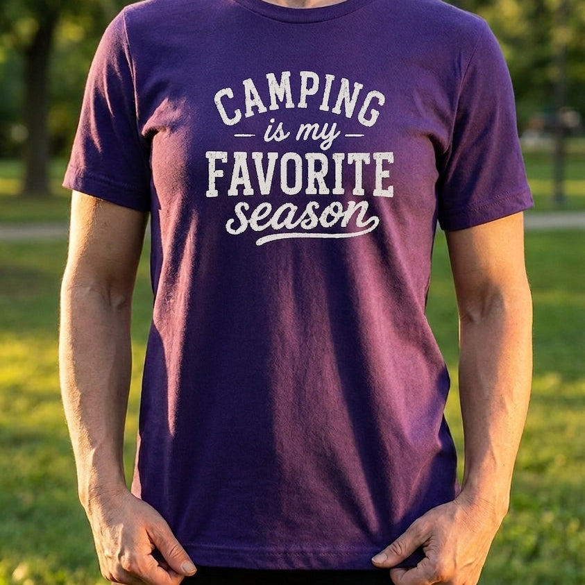 A woman wearing a purple short-sleeve t-shirt with the text 'CAMPING is my FAVORITE season' printed in white on the front, standing outdoors in a grassy park.