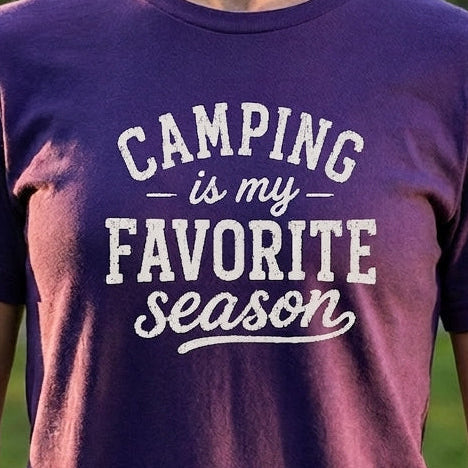 A woman wearing a purple short-sleeve t-shirt with the text 'CAMPING is my FAVORITE season' printed in white on the front, standing outdoors in a grassy park.