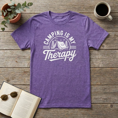 A heather purple short-sleeve t-shirt with a white graphic reading 'CAMPING IS MY Therapy' and showing a tent, trees, and stars, laid flat on a rustic wooden surface with a plant, coffee mug, book, and sunglasses as props.