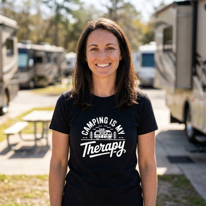 a woman with dark hair is standing outside in between multiple RVs. She is wearing a black t-shirt that says "camping is my therapy" with a camper in the middle of it.