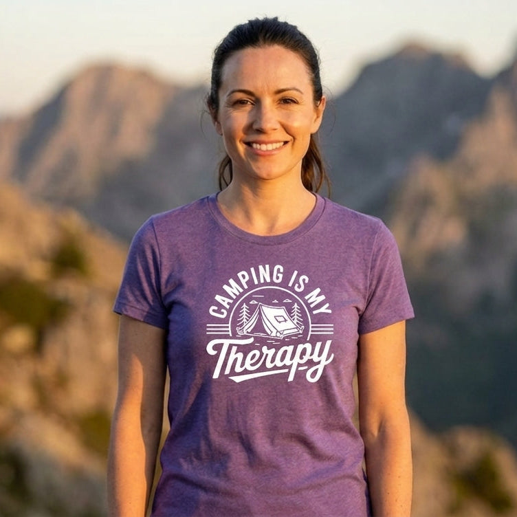 A woman smiling outdoors in front of mountains at sunset, wearing a purple short-sleeve T-shirt with a white graphic that reads 'CAMPING IS MY Therapy' and a camping tent illustration.