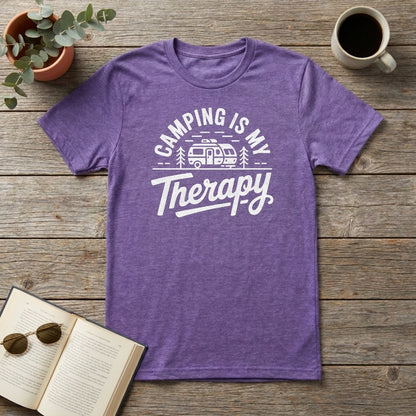 A heather purple short-sleeve t-shirt with a white graphic reading 'CAMPING IS MY Therapy' and a retro camper illustration, laid flat on a rustic wood surface with a plant, coffee mug, sunglasses, and open book as props.