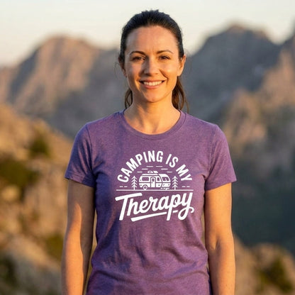 A woman smiling outdoors in front of mountains, wearing a purple short-sleeve T-shirt with white text that reads 'CAMPING IS MY Therapy' and a small camper graphic.