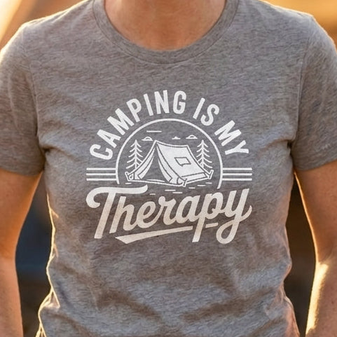 A woman smiling outdoors at sunset with a tent in the background, wearing a heather gray graphic T-shirt that reads 'CAMPING IS MY Therapy' in white lettering.