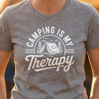 A woman smiling outdoors at sunset with a tent in the background, wearing a heather gray graphic T-shirt that reads 'CAMPING IS MY Therapy' in white lettering.