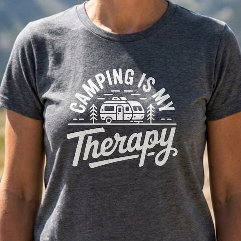 A smiling woman standing outdoors in a mountain landscape wearing a heather dark gray graphic T-shirt with white text that reads 'Camping is my Therapy' and a small camper illustration.