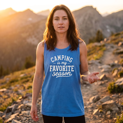 A woman standing outdoors on a mountain trail at sunset wearing a blue sleeveless tank top with the text 'CAMPING is my FAVORITE season' in white. The background shows rocky mountains and warm golden light.